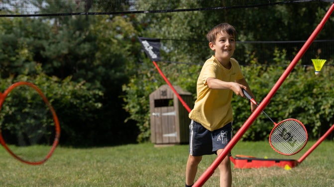 A boy in a yellow tshirt stands behind a net raised in the air. He is holding a badminton racket in his hand. A yellow shuttlecock is flying through the air
